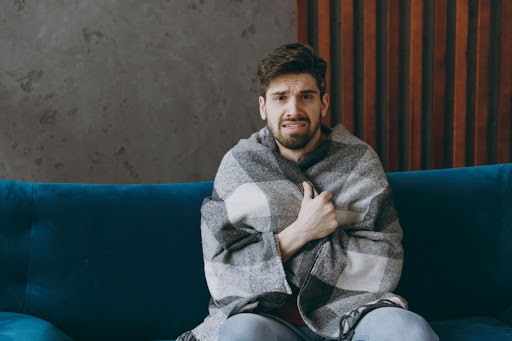 A man looking cold as he sits on a couch with a blanket.