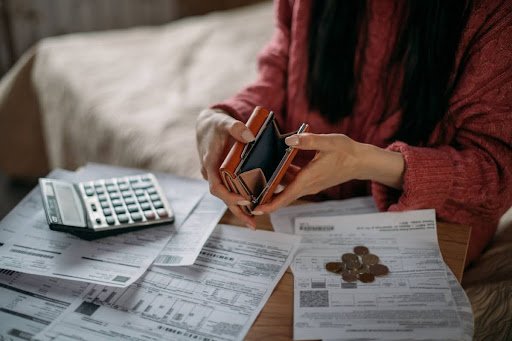 A woman opening a wallet with a calculator, bills, and coins on a table in front of her.