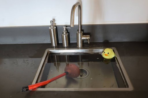 A clogged sink full of standing water with a rubber ducky and a plunger.