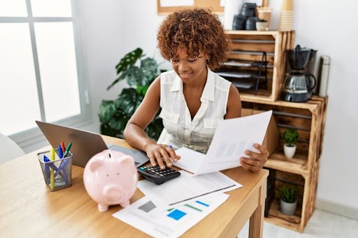 A woman using a calculator while she sits at a desk with a piggy bank, pens, papers, and a laptop.