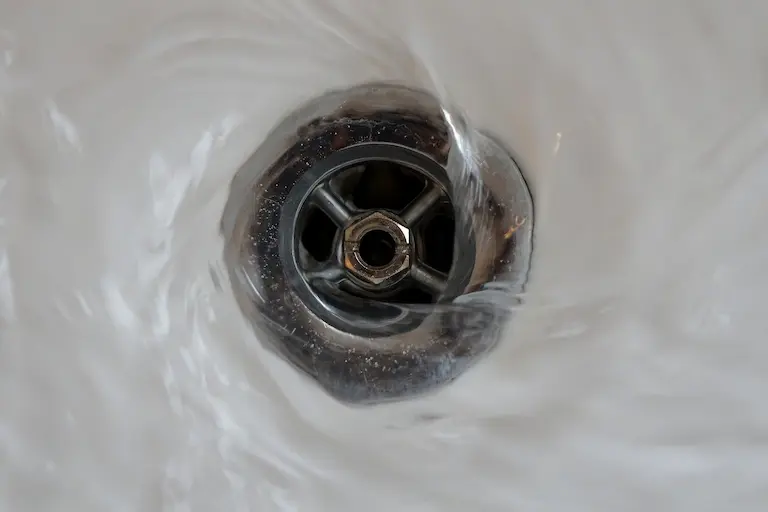 Close-up of water swirling and draining down a metal sink drain, creating a small whirlpool effect.