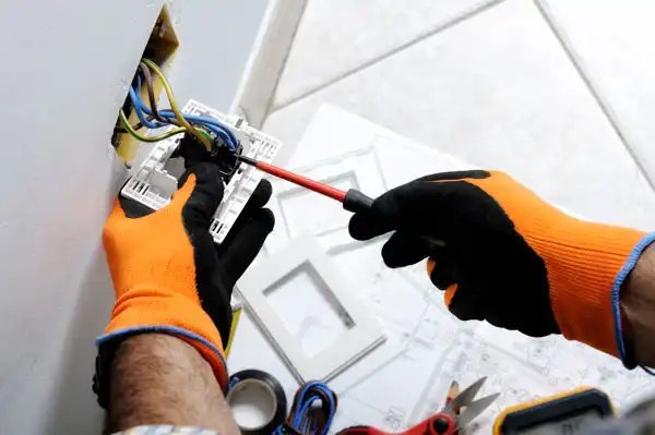 Person wearing orange gloves uses a screwdriver to install or repair electrical wiring in a wall socket. Tools and electrical tape are visible nearby.