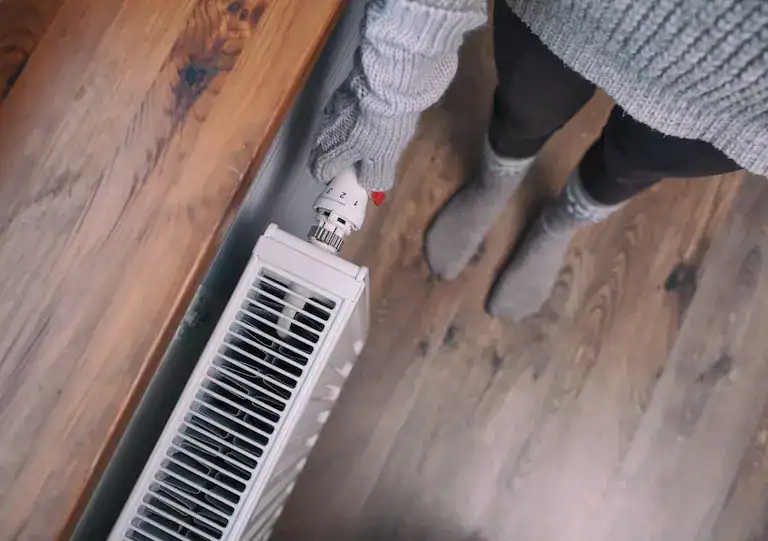 Person wearing a gray sweater and gloves adjusts the thermostat on a white radiator, standing on a wooden floor with gray socks visible.