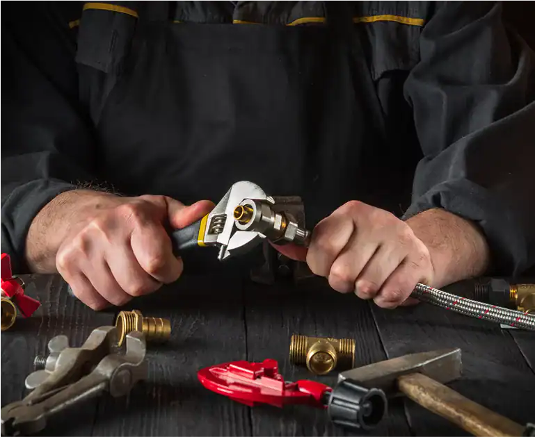 Person tightening a metal fitting with a wrench, surrounded by plumbing tools and pipe fittings on a dark wooden surface.