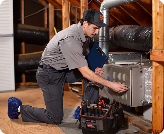 A technician kneels in an attic, inspecting a furnace with tools and a clipboard, while wearing protective shoe covers.